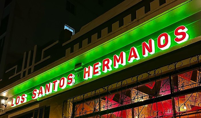 A fachada do Los Santos Hermanos à noite: o letreiro em neon verde e vermelho aceso sobre as janelas.